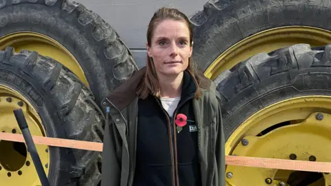 JOZEF HALL/BBC Cath Crowther looks to the right of the camera, she is wearing a green overcoat, black fleece, white top and with a poppy on her left side. She is standing in front of four tractor's wheels which are propped up against a wall. She has brown hair and blue eyes. 