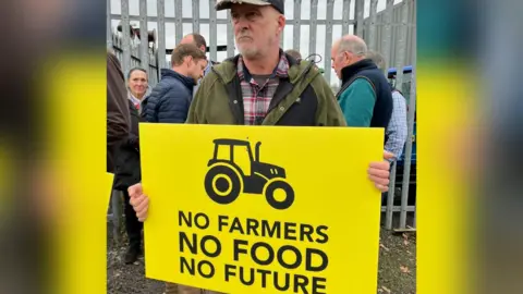 BBC A farmer holding a yellow banner with black writing saying No Farmers, No Food, No Future and a stylised image of a tractor stands outside Hexham Market.