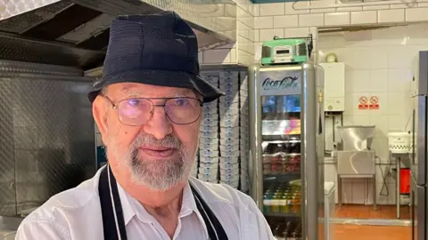 Annabel Amos/BBC John Panayis in a white shirt, a black apron and black hat stands in front of frying equipment.