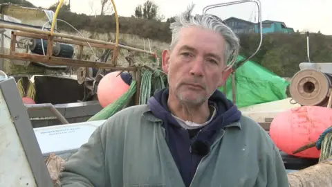 BBC Fisherman Stephen Andrews wearing a faded green overall looks at the camera. He is standing in front of a fishing boat.