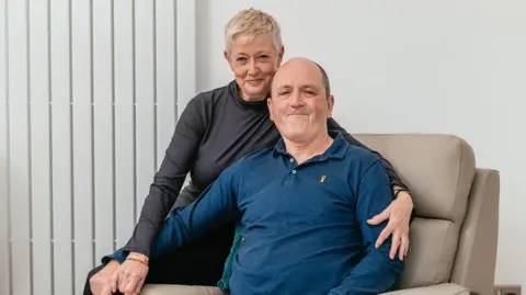 Owen Mathias/MNDA Mike Thomas, who has a bald head and is wearing a blue polo top, sat in a cream-coloured armchair and smiling while looking at the camera. Jayne who has short blonde hair and is wearing a black, long-sleeved turtle neck t shirt is sat behind him, on one of the arms of the chair, and has her arms round him