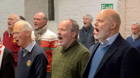 The Oxford Welsh Male Voice Choir singing. Some of the are wearing scarfs or tops with the Welsh flag.