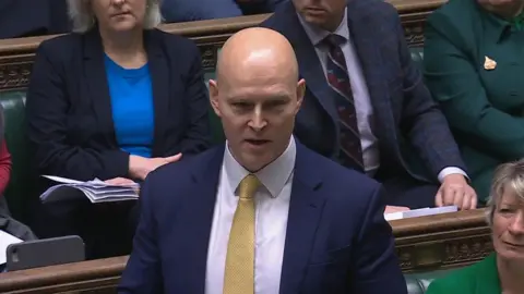 Parliament TV A screenshot of Max Wilkinson talking in Parliament this morning. He is bald with very fair eyebrows, and wears a dark suit with a white shirt and yellow tie. Other MPs can be seen behind him on the green leather benches. 