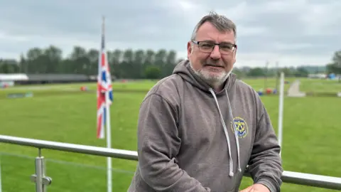 Rhea Alton Ian Bebbington is in a grey hoodie. He has grey hair and facial hair and is wearing black glasses. He is leaning on a metal rail of a balcony, behind him is a stretch of green grass and a white flagpole with the union jack hanging from the top.