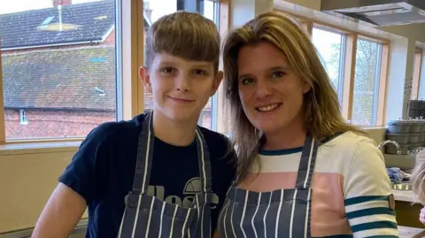 Ellen Roome Photograph of Ellen Roome and her son Jools. They are stood in a kitchen area with blue and white stripped aprons on.