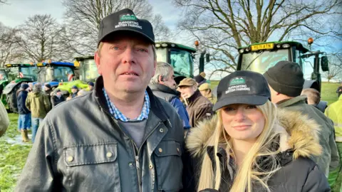 Linsey Smith/BBC Dave Adamson with his daughter Sarah. Both are wearing baseball-style caps with the words: "Supporting British farming" on them. Sarah has long blonde hair and is wearing a winter coat. Dave is wearing a wax jacket. There are tractors in the background.
