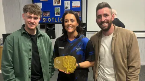 Pearson National Teaching Awards/Another Word Ciara Mulholland, who has dark hair, and is wearing a school tracksuit, stands in between singer ryan mcmullan and One Show presenter Ryan Hand. She is holding an award.