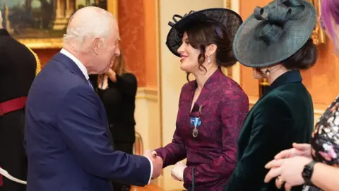 Getty Images The King is shaking hands with Georgia after presenting her with the medal at Buckingham Palace. The King is wearing a navy blue suit and is looking directly at Georgia and taking with her. Georgia is wearing a maroon coloured dress with a hat and is talking to the King. Her sister Melissa is stood next to her wearing a green dress and watching on.