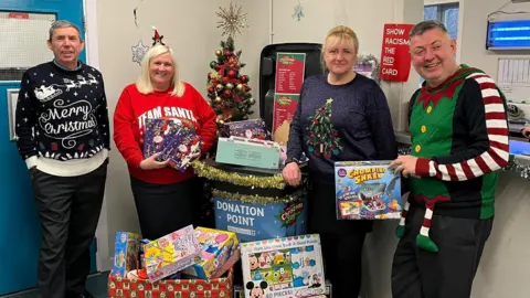Sefton Council Two men and two women wearing Christmas jumpers are stood at a bus depot with toys at a donation point.