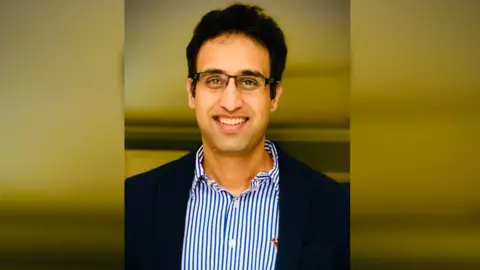 Dr Satpal Shekhawat A head and shoulders shot of a man wearing a blue and white striped shirt and a navy blazer. He has black hair and is wearing glasses.