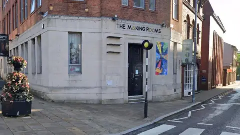 Google Street view image of the Making Rooms building in Blackburn. The ground floor is white brick and the top floor is red brick. It has long windows and a bright poster next to the black front door and stands next to a pedestrian crossing.