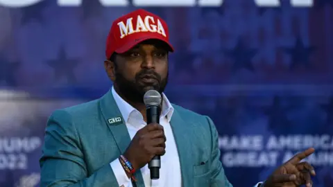 Getty Images Kash Patel, wearing a red MAGA hat, and speaking into a microphone and pointing
