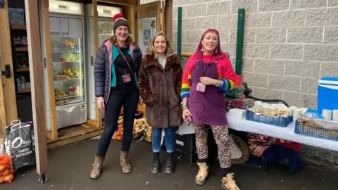 Shepton Mallet Community Fridge Three women including Vix Ross, right, stand in front of two fridges. They are all smiling, two women are wearing coats and Vix is wearing a bright pink jumper with a purple apron. A table of produce and drinks can be seen behind them