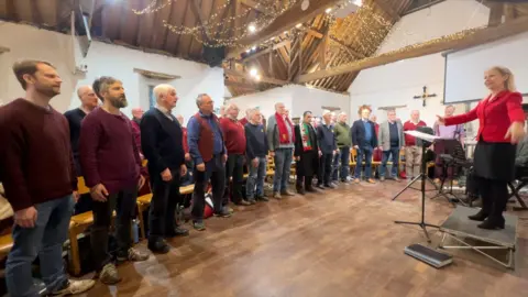 BBC The Welsh Male Voice Choir in Oxford singing in a room. A woman conducts them. There are lights on the ceiling and a crucifix.