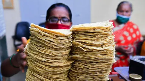 Getty Images In this photograph taken on March 8, 2021, members of Shri Mahila Griha Udyog, the organisation that produces the famous Lijjat Papad, weighs papadums at one of the organisation's facilities in Mumbai. - The fairytale success of Lijjat Papad -- a multi-million-dollar venture founded by seven women in a crowded Mumbai tenement in 1959 with seed capital of 80 rupees (1.10 USD) -- belies its revolutionary feminist aspirations. - TO GO WITH India-economy-gender-food, FEATURE by Ammu KANNAMPILLY (Photo by Indranil MUKHERJEE / AFP) / TO GO WITH India-economy-gender-food, FEATURE by Ammu KANNAMPILLY (Photo by INDRANIL MUKHERJEE/AFP via Getty Images)
