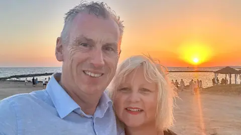 Family handout/PA Stuart Emerson standing on the beach beside his wife Claudia. He has short white hair and is wearing a light-blue collared shirt, smiling at the camera. Claudia has shoulder-length blonde hair and is wearing red lipstick, also smiling at the camera. Behind them the sun is setting on the horizon and the whole sky is lit up orange.
