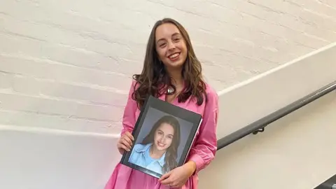 Chloe Morgan A woman, Chloe, stands on a flight of stairs holding a folder with a picture of her face on it. She is wearing a pink dress and smiling. 