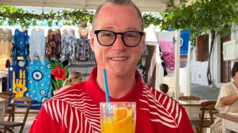 Facebook Man with short greying hair, smiling, looking at camera, wearing dark-rimmed glasses, wearing a red shirt with white pattern, holding a glass with orange liquid inside and an orange slice and blue straw, in what appears to be a cafe setting abroad, with bright towels and tops hanging up in the background.  