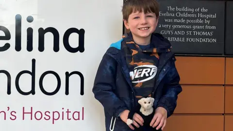 Guy's and St Thomas Trust Rufus Quinn is seen smiling while standing next to the Evelina Hospital sign holding a stuffed toy monkey.