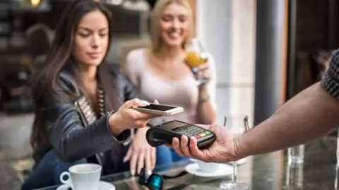 Getty Images A smiling woman with long dark hair and wearing a leather jacket uses her mobile phone to pay for a coffee in a café. Another woman with blond hair and wearing a white t-shirt is drinking a glass of orange juice.