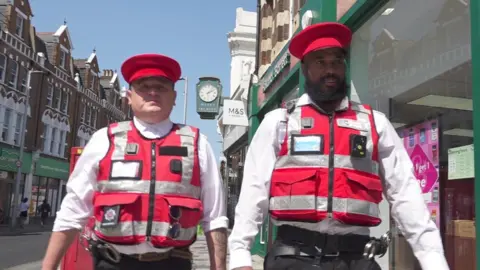BBC Two Bobbies, wearing red vests, red hats and white shirts, patrol on St John's Road in London.
