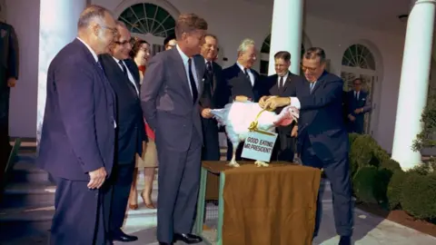 John F. Kennedy Presidential Library/NARA JFK stands with administration officials while a turkey is presented saying 'Good Eating Mr President' around its neck