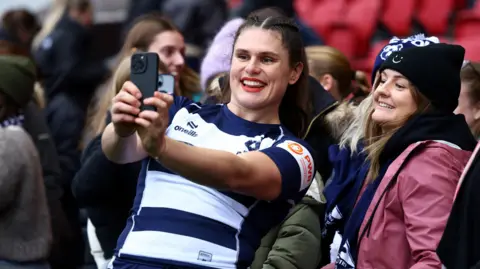 PA Media Bristol Bears' Ilona Maher takes a selfie with fans after the match with Gloucester/Hartbury at Ashton Gate. She is smiling as she holds the phone out to take the picture, as are the female fans in the background