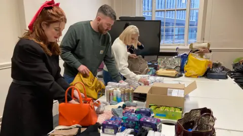 BBC Four people, a woman with ginger hair, a man with brown hair, a woman with blonde hair and a woman with black hair are standing over a table. The table has boxes of sanitary pads, gloves, body spray and handbags. They are packing the handbags with the items.