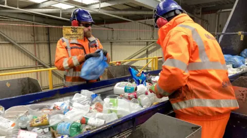 BBC Two men in high vis uniform standing by a conveyor belt sorting mixed plastics