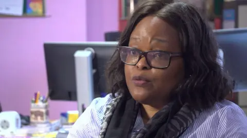 Jacqui Mukono, who sits looking slightly off camera during TV interview. She is wearing a striped blue and white shirt, a black scarf and black glasses.