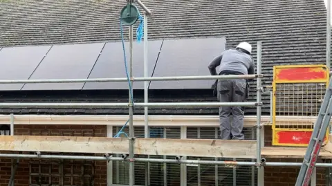 BBC A man dressed in grey with a white hard hat is turned away from the camera, standing on scaffolding and working on a solar panel, which is fixed to the roof of a bungalow. 