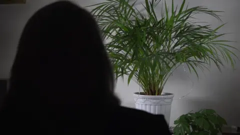 A silhouette of a woman in a darkened room with a pot plant in the background.