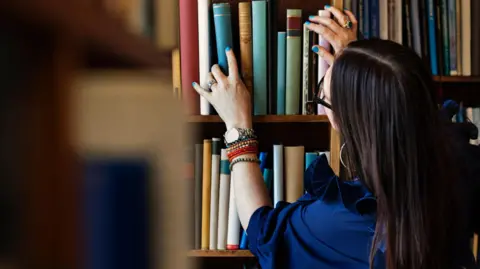 Getty Images A stock image of a library worker, looking at books. She has long dark hair, is wearing a blue top, and has on a watch and bracelets on her left arm. She has a ring on her right hand, which is raised and on a book. The are a large number of books on shelves.