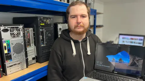 BBC Billy holds a laptop PC showing the Windows home screen as he stands in a workshop in front of shelves full of desktop computers. He has ginger hair and a short beard and is wearing a black hoodie.