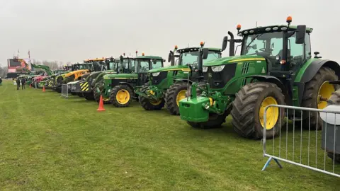 Richard Knights/BBC A long line of green tractors parked on grass with some orange bollards nearly and people at the end of the field. The sky is grey.