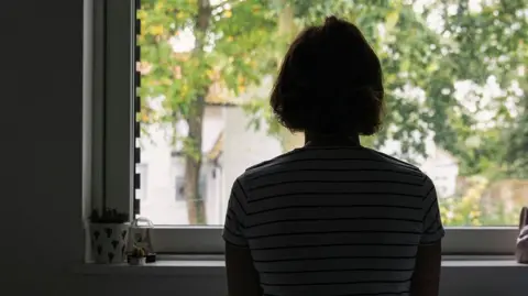Getty Images A woman sat in front of a window with her back to the camera. She has bobbed hair and is wearing a striped t shirt