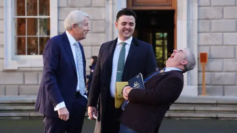 PA Media Members of Regional Independent Group (left to right) Michael Lowry, Barry Heneghan and Kevin "Boxer" Moran at Leinster House in Dublin. All are wearing suits, Kevin "Boxer" Moran is bent over laughing, with the others smiling at him.