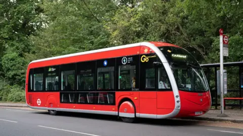 TfL File image of one of the new 358 red single-decker buses, which have a slanted windscreen that curves under at the bonnet, lower windows for some seats, and red hubcaps over the tyres. The pantograph arm is not visible