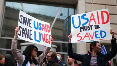 Getty Images People holding protest signs in black, blue and red, reading: 'Save USAID, save lives' and 'USAID must be saved'