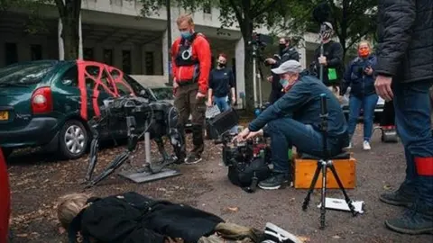 Fox TV An actor lies on the floor in a chaotic street scene, with the camera operator and crew wearing masks