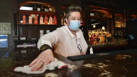 PAcemaker A female bar worker with short hair, wearing a disposable face covering, standing behind a bar and cleaning down the bar counter