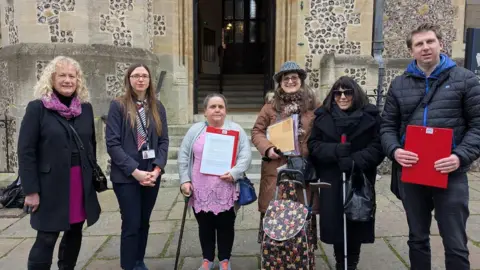 Waterside Changemakers A group of six people stand in front of a stone-walled building holding clipboards containing signatures on a petition. The group is made up of 5 women and one man. The two people on the left of the image are Councillor Lulu Bowerman and Councillor Kirsty North.