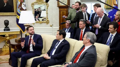 Reuters US Vice President JD Vance, sitting on a sofa in the Oval Office, speaks to a group of reporters behind him holding microphones.