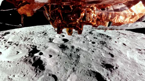 Reuters The Moon passes underneath the Blue Ghost spacecraft on 24 February 2025 as it orbits the lunar surface ahead of its landing on 2 March 2025. The gold foil underneath the spacecraft is visible, below is the cratered gray surface of the moon and the blackness of space is in the background.