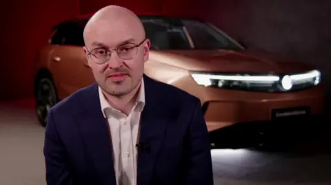 A man with a shaven head, glasses and a short, stubbly beard, and wearing a suit, sits in front of a large screen or wall with a picture of an orange electric car covering the whole background space