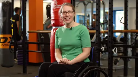 PA Media Claire Trivedi, a woman with long blonde hair and glasses wearing a green T-shirt and black trousers, sits in a wheelchair in a gym. Various equipment including punchbags and wall-mounted bars can be seen behind her.