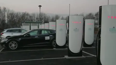 BBC A row of charging bays in a car park, with white towers installed in every parking space. A black electric car and a white electric car are both parked up and charging.