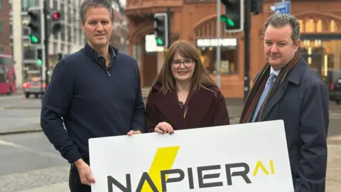 BBC Napier AI's CEO Greg Watson, Economy Minister Caoimhe Archibald and Invest NI CEO Kieran Donoghue stand holding a large white sign with NapierAI written on it. Mr Watson is wearing a navy quarter-zip jumper, Ms Archibald is wearing a burgundy coat and Mr Donoghue is wearing a navy coat and a striped scarf.