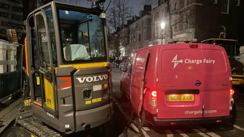 Transport for London A Volvo branded, grey and yellow construction vehicle being charged by a red van with the branding 'Charge Fairy' on a road.