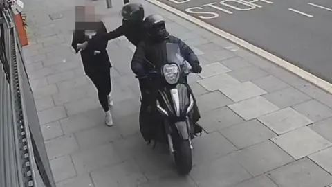 Metropolitan Police Two people on a moped are driving on the pavement past a pedestrian and one is reaching out to snatch the person's phone from their hand